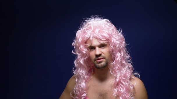 Сумасшедший красивый молодой парень в кудрявом парике и розовой футболке на темном фоне танцует смешно, показывает свои мускулы — стоковое видео