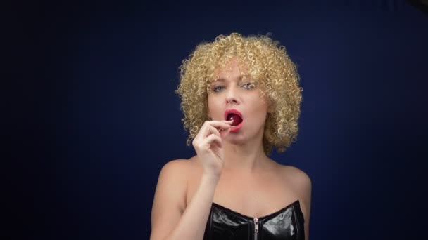Sladká žena s krátkými kudrnatými blond vlasy v černém koženém korzetu olizuje červené lesklé lízátko. tmavé pozadí — Stock video
