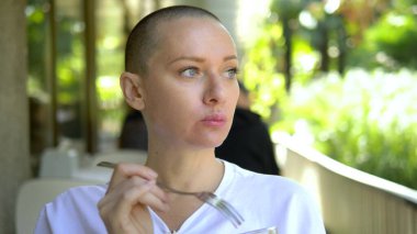 Güzel, genç, kel bir kadın açık bir kafede yemek yiyor..