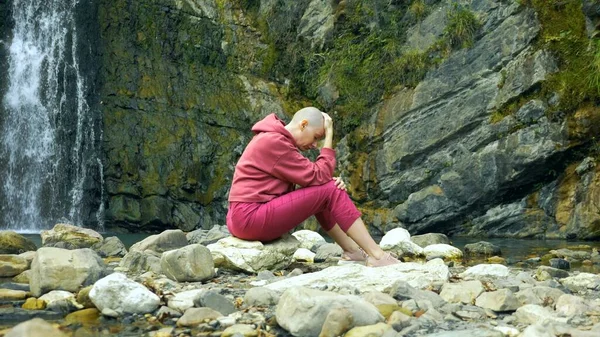 Sad bald woman sits on a stone against the background of a waterfall. copy space. concept of unity with nature, hiding from people.