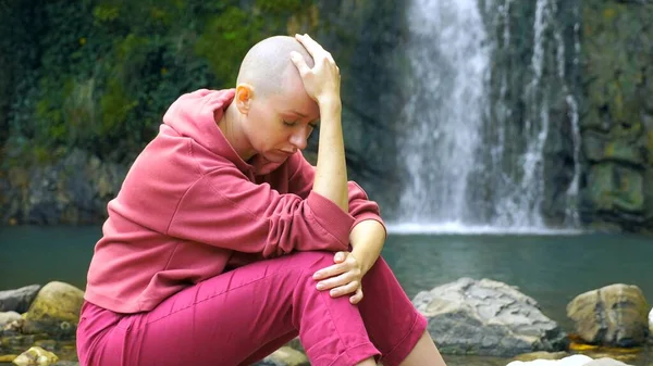 Wanita botak sedih duduk di atas batu dengan latar belakang air terjun. Salin ruang. konsep persatuan dengan alam, bersembunyi dari orang-orang . — Stok Foto