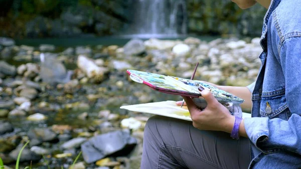 Primer plano. Una artista. manos en la naturaleza contra el telón de fondo de una cascada. pintura en la paleta — Foto de Stock