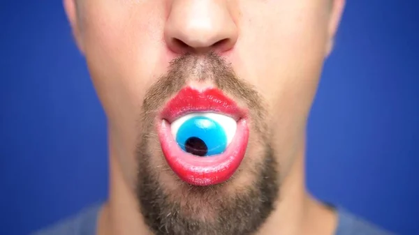 Zbliżenie. Brodaty mężczyzna z pomalowanymi ustami trzyma niebieskie oko cukierka w ustach. — Zdjęcie stockowe