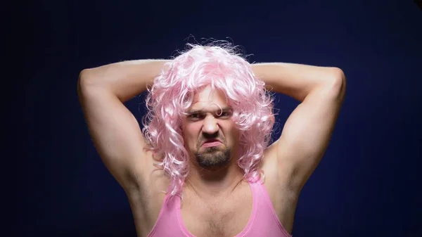 Pazzo bel ragazzo con una parrucca riccia e una t-shirt rosa su uno sfondo scuro sta ballando divertente, mostra i suoi muscoli — Foto Stock