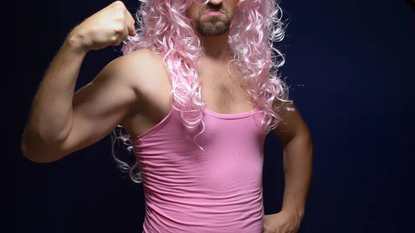 Loco guapo joven en una peluca rizada y una camiseta rosa sobre un fondo oscuro está bailando divertido, muestra sus músculos —  Fotos de Stock