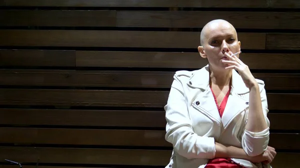 Bald girl in a leather white jacket smokes and drinks on the terrace in a nightclub — Stock Photo, Image