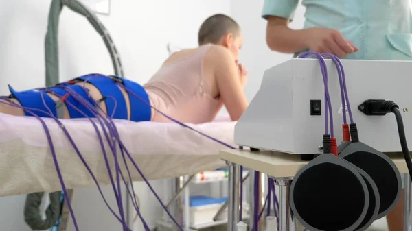 A menina se deita no sofá e recebe o procedimento de mioestimulação em suas pernas e nádegas em um salão de beleza. Cuidado corporal, perda de peso . — Fotografia de Stock