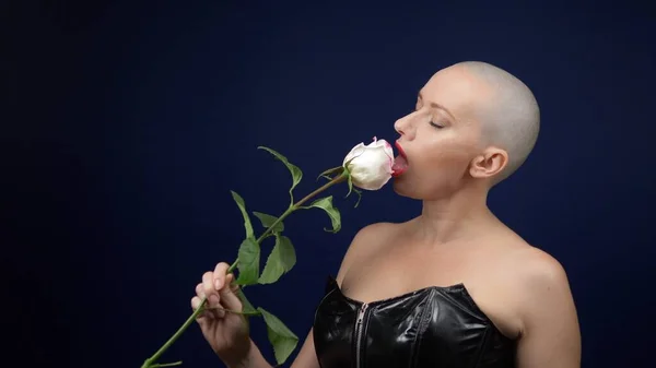 Die schöne, glatzköpfige Frau im schwarzen Lederkorsett hält eine rosa Rose in den Händen und küsst eine Blume. Abenteuer fremder Menschen. — Stockfoto