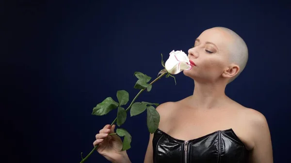 Όμορφη φαλακρή γυναίκα με μαύρο δερμάτινο κορσέ κρατά ένα ροζ τριαντάφυλλο στα χέρια της και φιλάει ένα λουλούδι. περιπέτειες παράξενων ανθρώπων. — Φωτογραφία Αρχείου