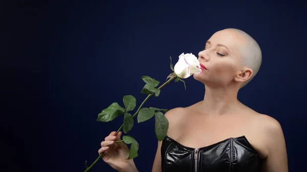 Die schöne, glatzköpfige Frau im schwarzen Lederkorsett hält eine rosa Rose in den Händen und küsst eine Blume. Abenteuer fremder Menschen. — Stockfoto