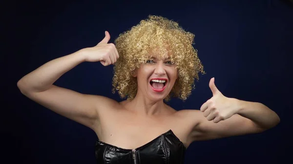 Una mujer traviesa con el pelo rizado blanco corto en un corsé de cuero negro muestra un signo estupendo, pulgares hacia arriba. emociones y gestos. espacio de copia — Foto de Stock