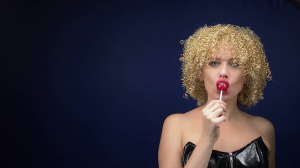 Милая женщина с короткими вьющимися светлыми волосами в черном кожаном корсете лижет красный блестящий леденец. темный фон

