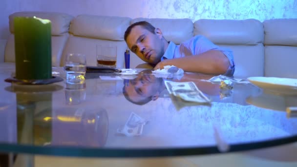 Молодий чоловік відчуває галюцинації від прийому наркотиків і алкоголю, лежачи на підлозі поруч зі столом у вітальні . — стокове відео