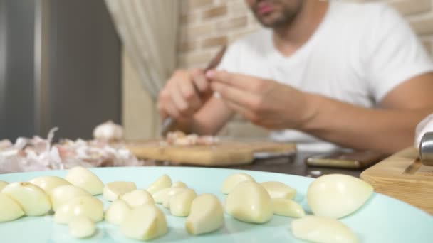 Een man schilt knoflook zittend aan een tafel op een achtergrond van een bakstenen muur. — Stockvideo