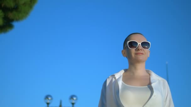 Elegante ragazza calva in occhiali da sole e una camicia bianca cammina per strada contro un cielo blu e alberi verdi — Video Stock