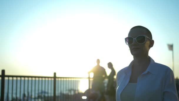 Stylish bald girl in sunglasses and a white shirt walks the street against a blue sky and green trees — Stock Video