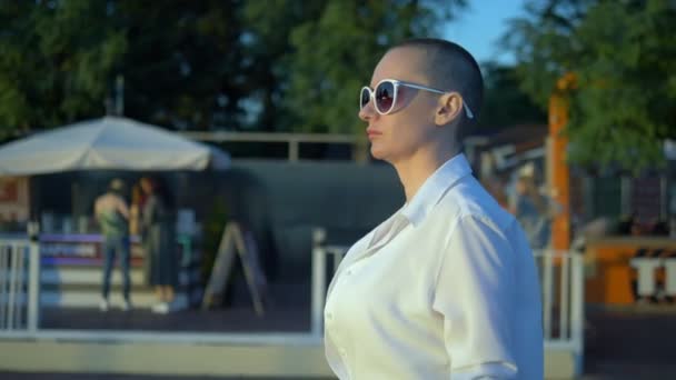 Stylová holohlavá dívka v slunečních brýlích a bílé košili kráčí ulicí proti modré obloze a zeleným stromům — Stock video