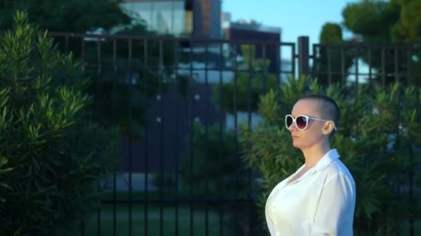 Stilvolles glatzköpfiges Mädchen mit Sonnenbrille und weißem Hemd geht die Straße vor blauem Himmel und grünen Bäumen entlang — Stockvideo