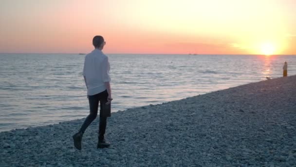 Jong mooi kaal meisje wandelend op het strand bij zonsondergang. Een vrouw in een wit shirt, leren broek en laarzen loopt langs de kust genietend van de zonsondergang. — Stockvideo