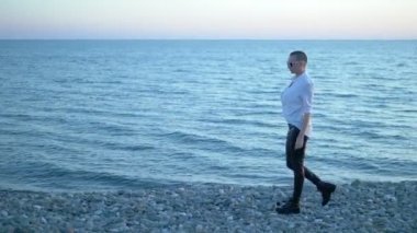 Gün batımında sahilde yürüyen güzel kel kız. Beyaz gömlekli, deri pantolonlu ve botlu bir kadın deniz kenarında günbatımının tadını çıkarıyor..