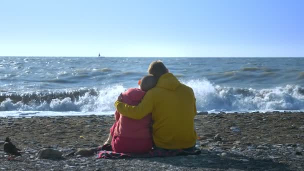 Pareja en el hombre del amor y una mujer calva están sentados en la orilla del mar en tiempo ventoso fresco . — Vídeos de Stock