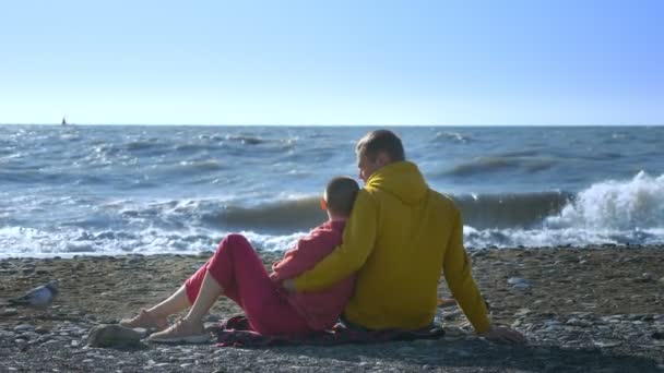 Pareja en el hombre del amor y una mujer calva están sentados en la orilla del mar en tiempo ventoso fresco . — Vídeos de Stock