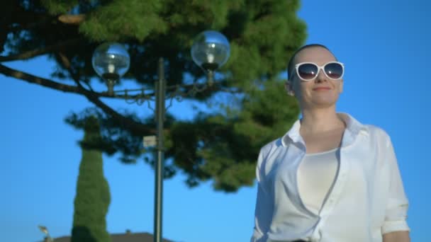 Κομψό φαλακρό κορίτσι με γυαλιά ηλίου και ένα λευκό πουκάμισο περπατά στο δρόμο ενάντια σε ένα μπλε ουρανό και πράσινα δέντρα — Αρχείο Βίντεο