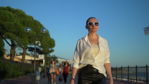 Chica calva con estilo en gafas de sol y una camisa blanca camina por la calle contra un cielo azul y árboles verdes — Vídeos de Stock