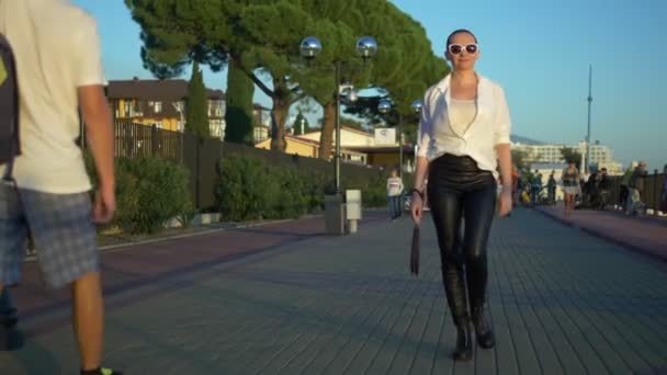 Stijlvol kaal meisje in zonnebril en een wit shirt loopt de straat tegen een blauwe hemel en groene bomen — Stockvideo