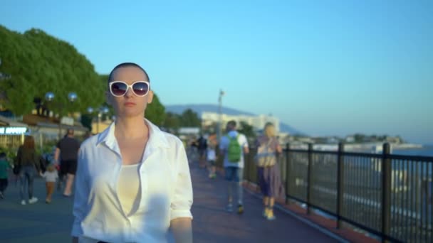 Menina careca elegante em óculos de sol e uma camisa branca caminha na rua contra um céu azul e árvores verdes — Vídeo de Stock