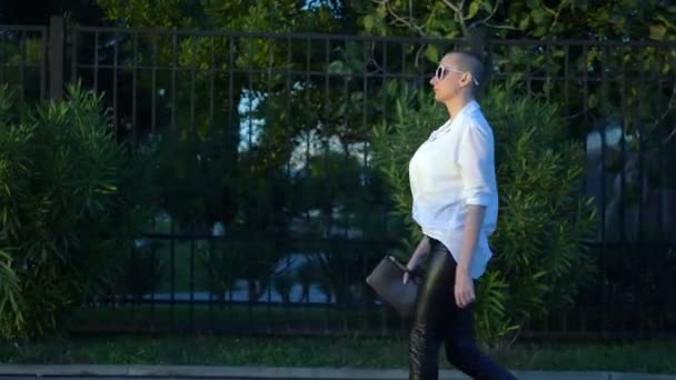 Стильная лысая девушка в солнечных очках и белой рубашке идет по улице против голубого неба и зеленых деревьев — стоковое видео