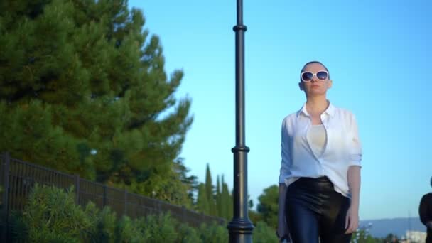 Stilvolles glatzköpfiges Mädchen mit Sonnenbrille und weißem Hemd geht die Straße vor blauem Himmel und grünen Bäumen entlang — Stockvideo