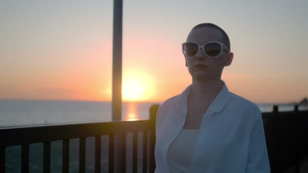 Joven chica calva hermosa caminando en la playa al atardecer. Una mujer con una camisa blanca, pantalones de cuero y botas camina por la orilla del mar disfrutando de la puesta de sol . — Vídeos de Stock