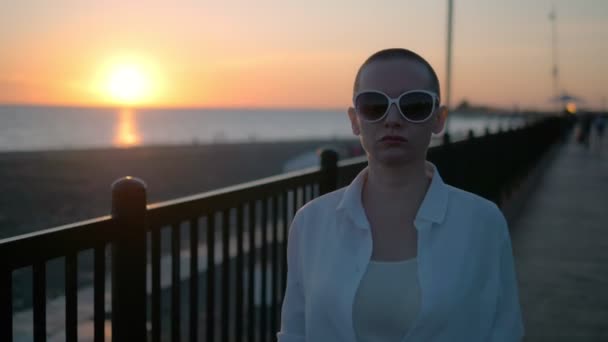 Joven chica calva hermosa caminando en la playa al atardecer. Una mujer con una camisa blanca, pantalones de cuero y botas camina por la orilla del mar disfrutando de la puesta de sol . — Vídeo de stock