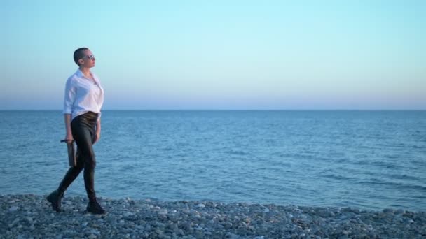 Joven chica calva hermosa caminando en la playa al atardecer. Una mujer con una camisa blanca, pantalones de cuero y botas camina por la orilla del mar disfrutando de la puesta de sol . — Vídeo de stock