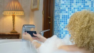 Sarışın kız musluk suyunun altında akıllı telefonu yıkıyor ve lüks bir banyoda köpükle banyo yapıyor. Esprili ya da su geçirmez telefon kavramı.