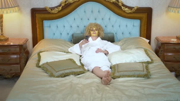 Jonge blonde vrouw met een gouden masker op haar gezicht tv kijken liggend op een luxe gouden bed. — Stockvideo