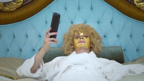 Young blond woman with a golden mask on her face watching television lying on a luxurious golden bed. — Stock Video