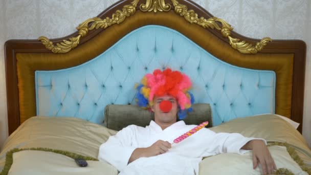 A young man in a white coat and a clown wig lies on a luxurious golden bed and eats a lollipop on a stick. concept of humor, adventure of strange people. — Stockvideo