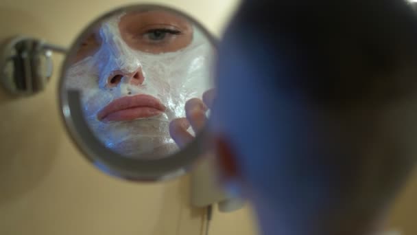 Chica calva en un abrigo blanco pone una máscara de belleza en su cara, mirando en el espejo en el baño . — Vídeos de Stock