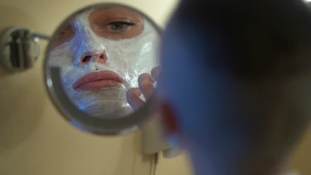 Bald girl in a white coat puts a beauty mask on her face, looking in the mirror in the bathroom. — Stockvideo