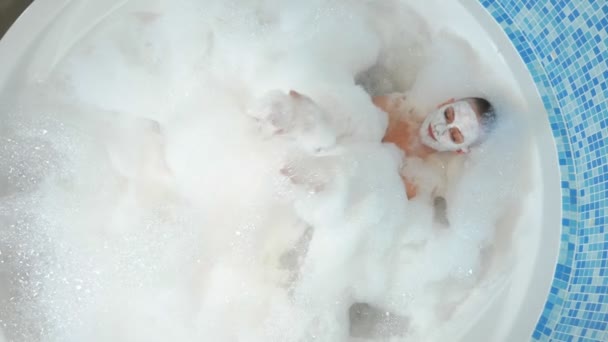 View from above. An attractive young bald woman with a mask on her face lies in a bathtub with thick lush foam. rest, relaxation — Stock Video