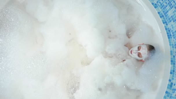 Vista desde arriba. Una atractiva joven calva con una máscara en la cara yace en una bañera con espuma espesa y exuberante. descanso, relajación — Vídeo de stock