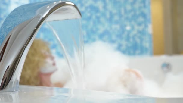 V ohniskové vodě tekoucí z kohoutku do vany, na rozmazaném pozadí Mladá žena v masce si dá bublinkovou koupel — Stock video