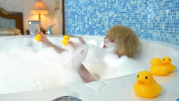 Schöne junge blonde Frau mit einer Maske auf dem Gesicht beim Baden mit Blasen spielt mit einer gelben Ente. Humorvolles Konzept — Stockvideo