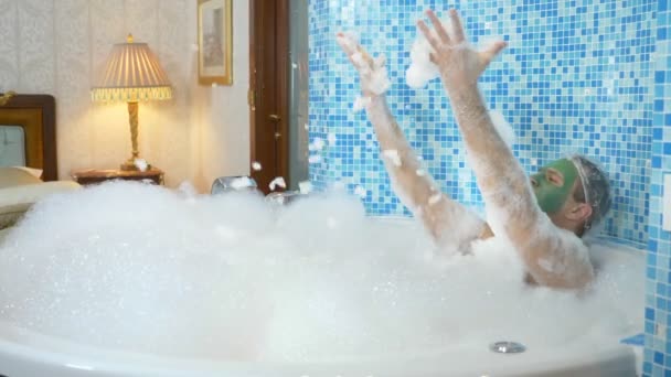 Молодий чоловік з зеленою маскою на обличчі і в душовій шапці приймає ванну з піною. хлопець танцює у ванній кімнаті, його тіло покривається густою білою піною. копіювання простору — стокове відео