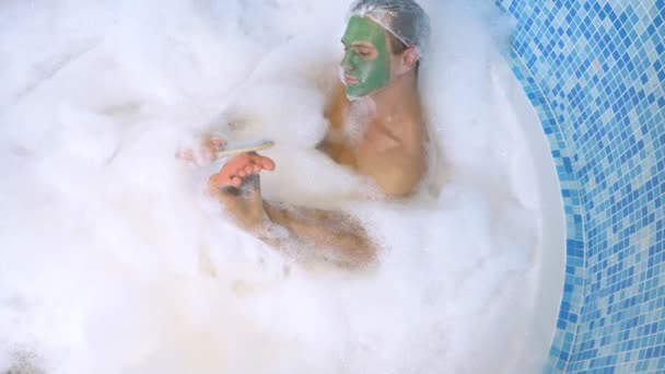 A man in a shower cap and a green face mask lies in a bath with copious foam and washes his foot with pumice. hygiene with scrub in the bath. view from above — 비디오