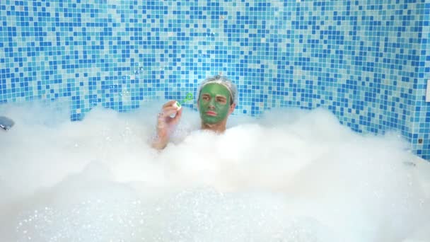 Homem bonito emocional com máscara de barro em uma touca de banho, soprando bolhas de sabão enquanto deitado em uma banheira com espuma abundante. conceito humorístico. espaço de cópia — Vídeo de Stock
