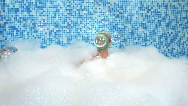 Homme mignon émotionnel avec masque en argile dans un bonnet de bain, soufflant des bulles de savon tout en étant couché dans une baignoire avec une mousse abondante. concept humoristique. espace de copie — Video