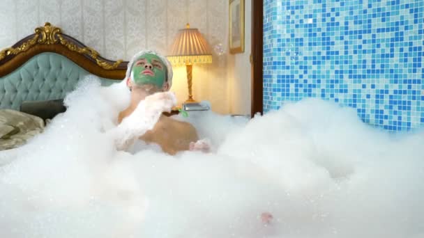 Емоційний милий чоловік з глиняною маскою в шапці для купання, що дме мильні бульбашки, що лежать у ванні з багатою піною в розкішній ванній кімнаті. копіювання простору — стокове відео
